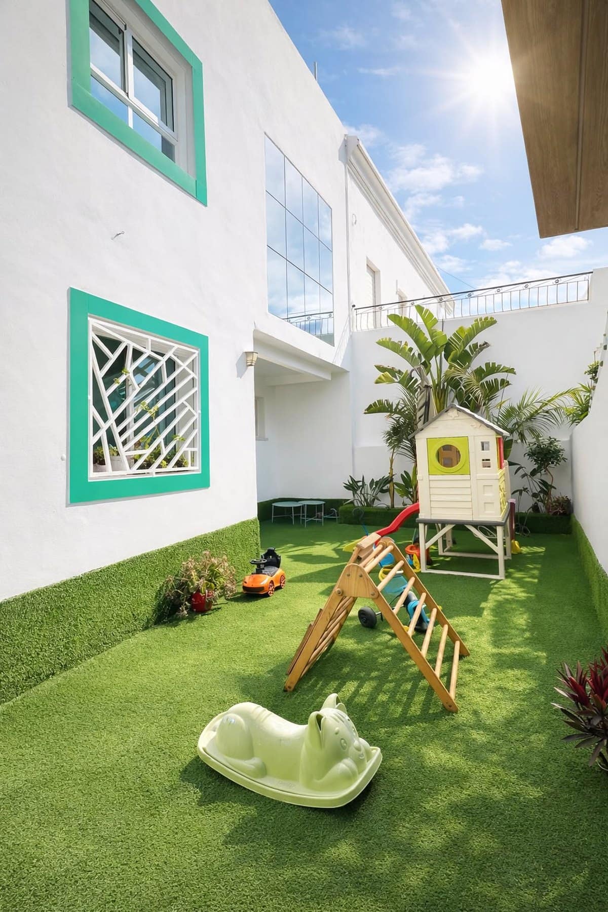 Le jardin de la crèche : gazon, maisonnette blanche avec toboggan et triangle de motricité en bois, sous le soleil